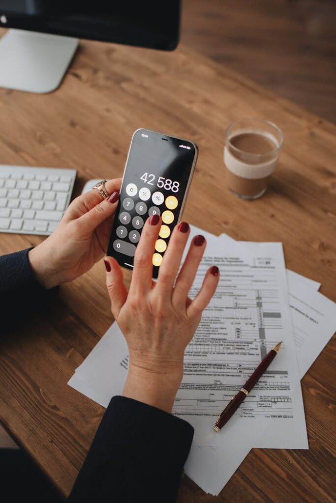 Hands using smartphone calculator over documents on a wooden table, indicating financial calculation or budgeting.